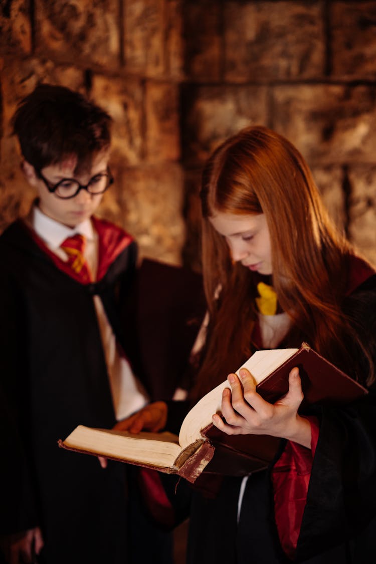 A Boy And Girl Standing In Black Robes Reading A Spell Book