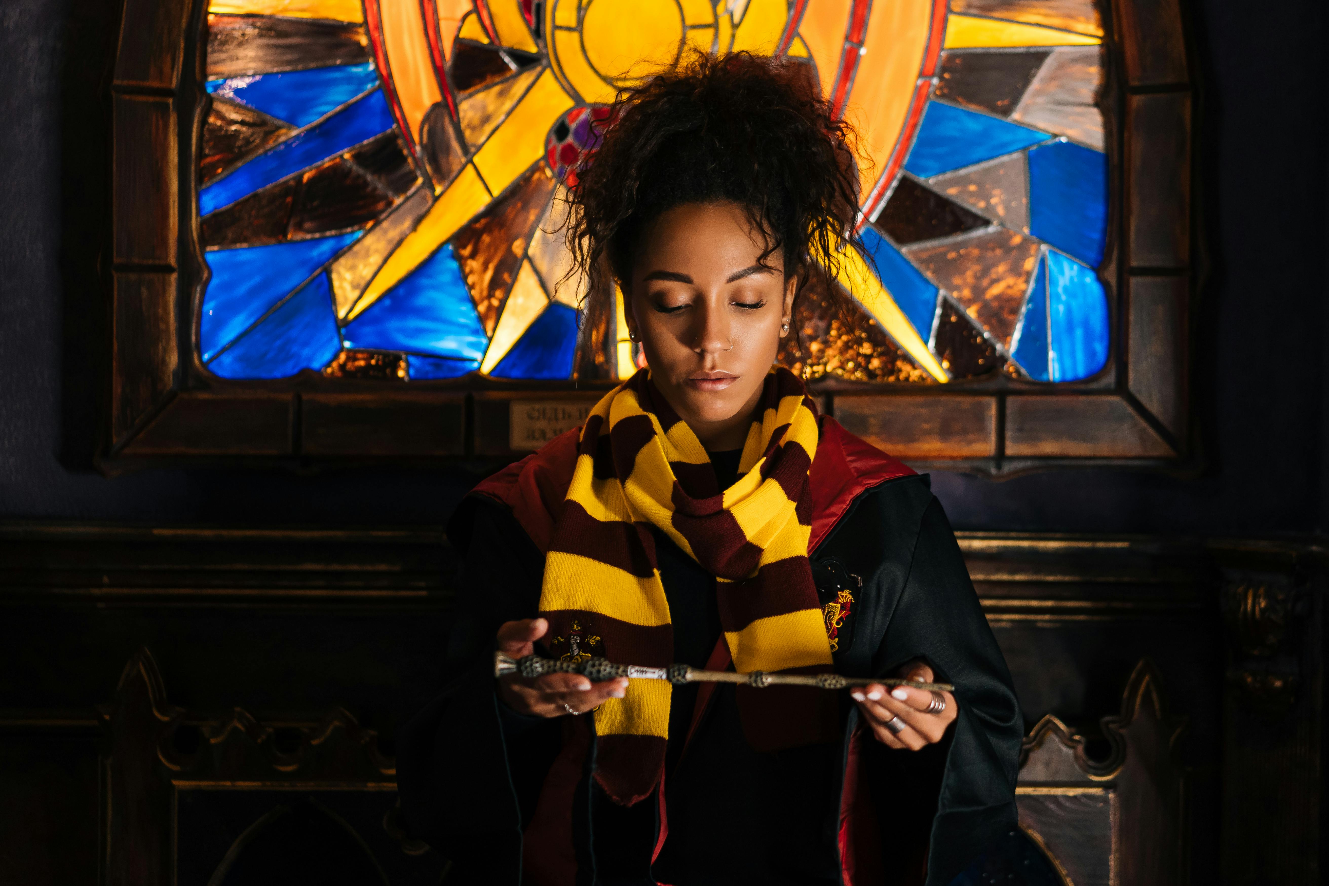 A young woman dressed as a wizard holding a wand in front of a stained glass window.