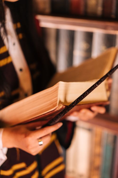 Free A woman in a Harry Potter-inspired costume holding an open book in front of a bookcase. Stock Photo