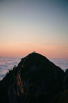Hikers silhouetted against a colorful sunset atop a mountain peak, surrounded by clouds.