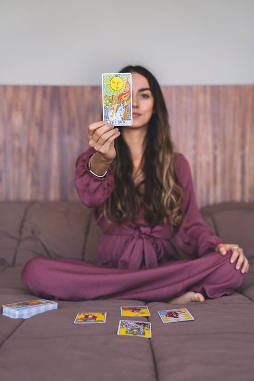 Free A woman sitting indoors in a purple dress holding up a tarot card, suggesting a spiritual reading. Stock Photo