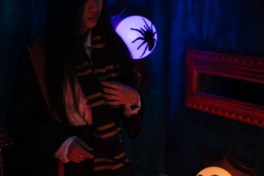A woman dressed as a witch in a dark setting, holding a wand and scarf, illuminated by a spider lamp.