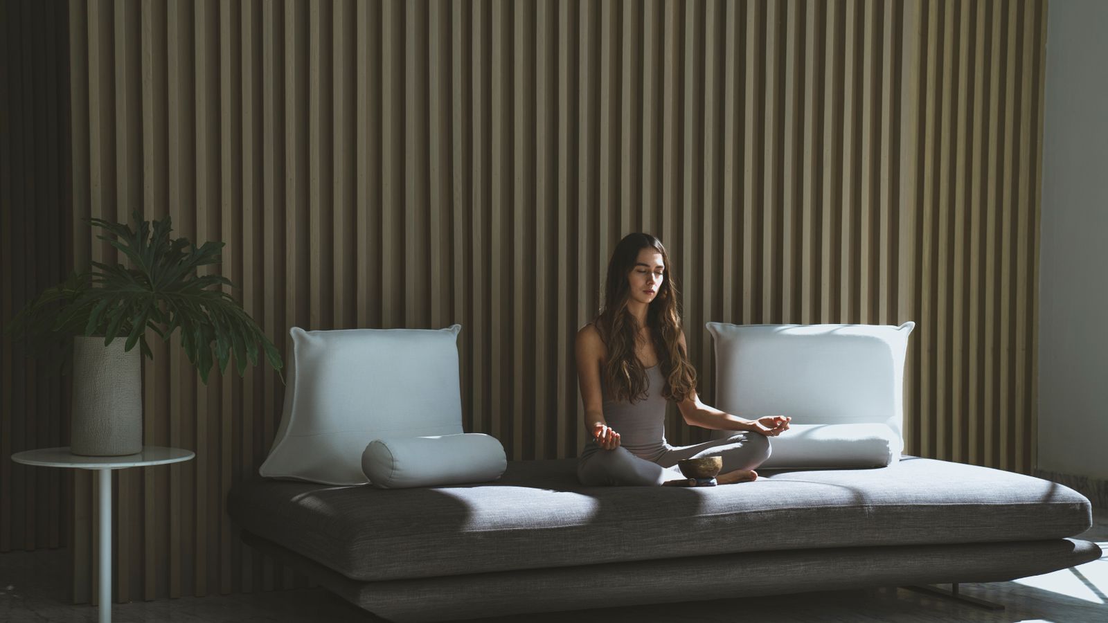 A woman meditating with her eyes closed while sitting on a gray sofa indoors