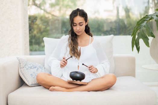 A young woman in white meditates indoors while playing a steel tongue drum, creating a serene atmosphere.