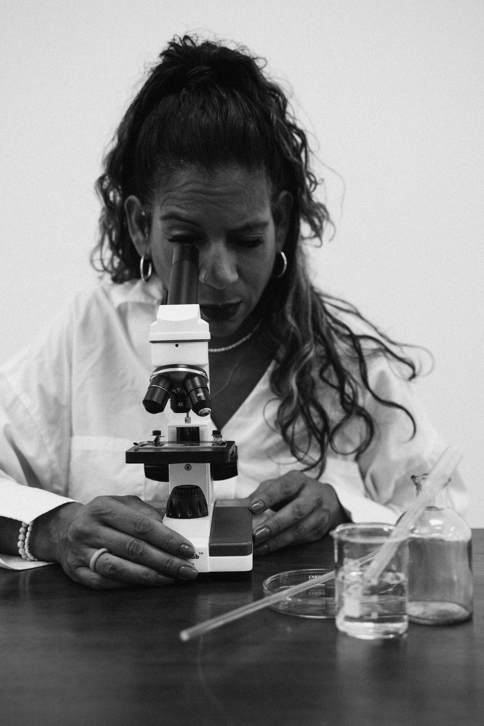 Black and white photo of a woman scientist in a lab coat looking through a microscope beside laboratory glassware