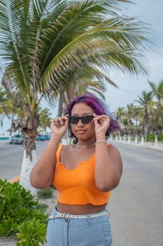 Young woman with colored hair adjusting sunglasses under palm trees on a sunny day street.