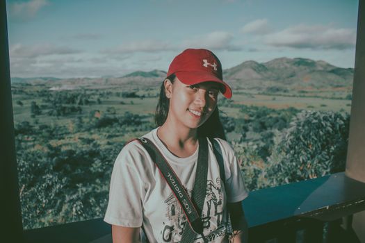 A young woman wearing a red cap smiles with a scenic mountain landscape in the background.