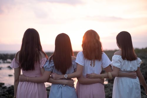 Free Four women stand arm in arm, facing a serene sunset at the beach. Stock Photo