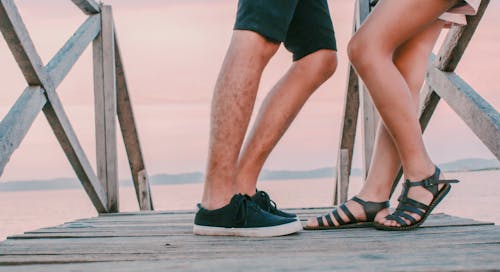 Man in Black Low-top Sneakers Leaning Towards Woman in Black Gladiator Flat Sandals on Dock