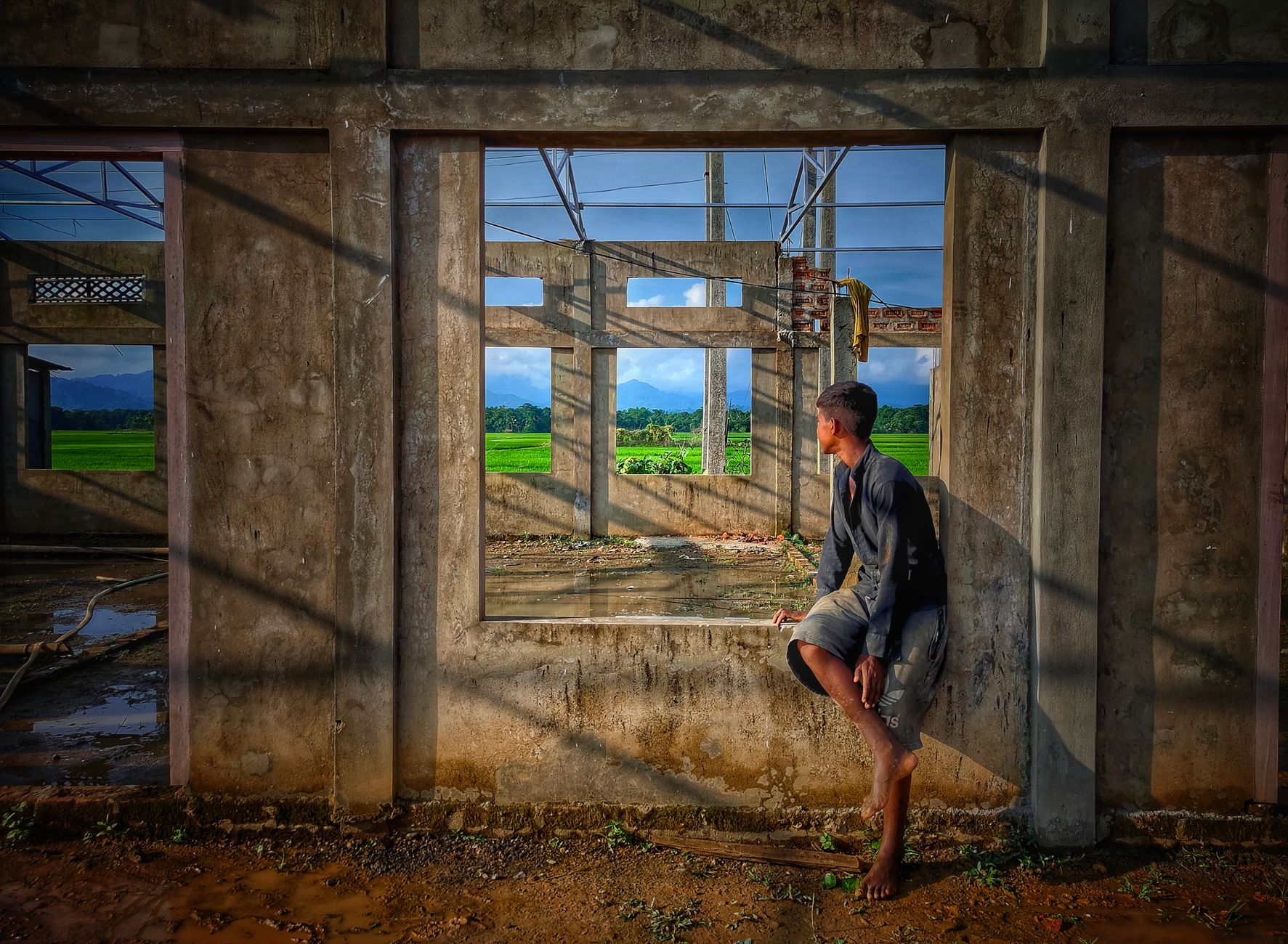 A young man in casual attire sits in an unfinished building, looking out at a lush landscape through a window frame.