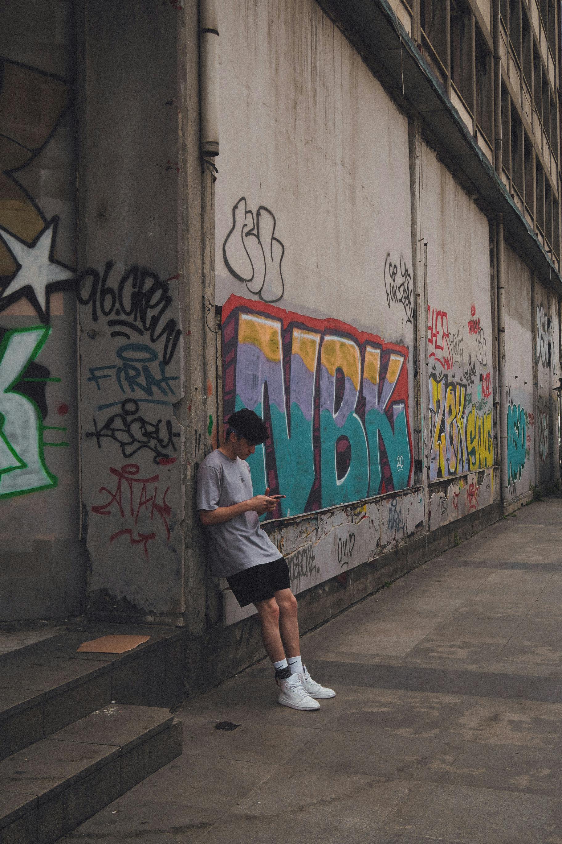trendy young male millennial using smartphone leaning on graffiti wall