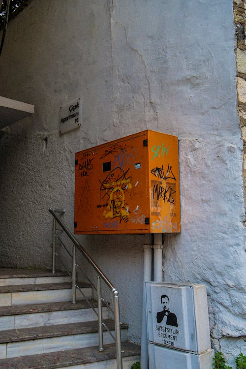Free Aged orange metal power box with inscriptions and graffiti hanging on white concrete wall above stairs of aged building in city Stock Photo