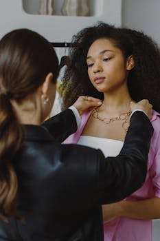 A stylist assists a woman trying on a gold necklace in an indoor fashion store setting.