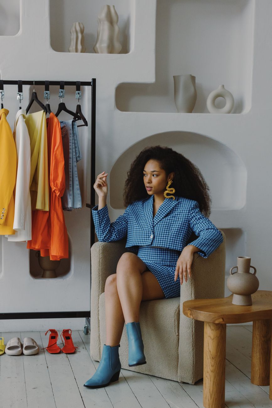 Stylish black woman in a blue blazer seated near colorful clothes in a modern room.
