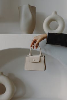 Elegant beige handbag held by a hand, surrounded by modern sculptures on a neutral background.