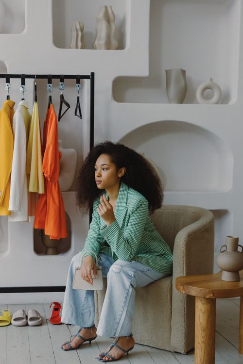 Free Fashionable woman with afro hair in a green jacket seated on a chair with colorful clothes in the background. Stock Photo