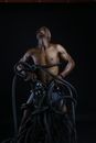 Serious young shirtless African American muscular man yelling with close eyes while trying to break free from black ropes in dark studio