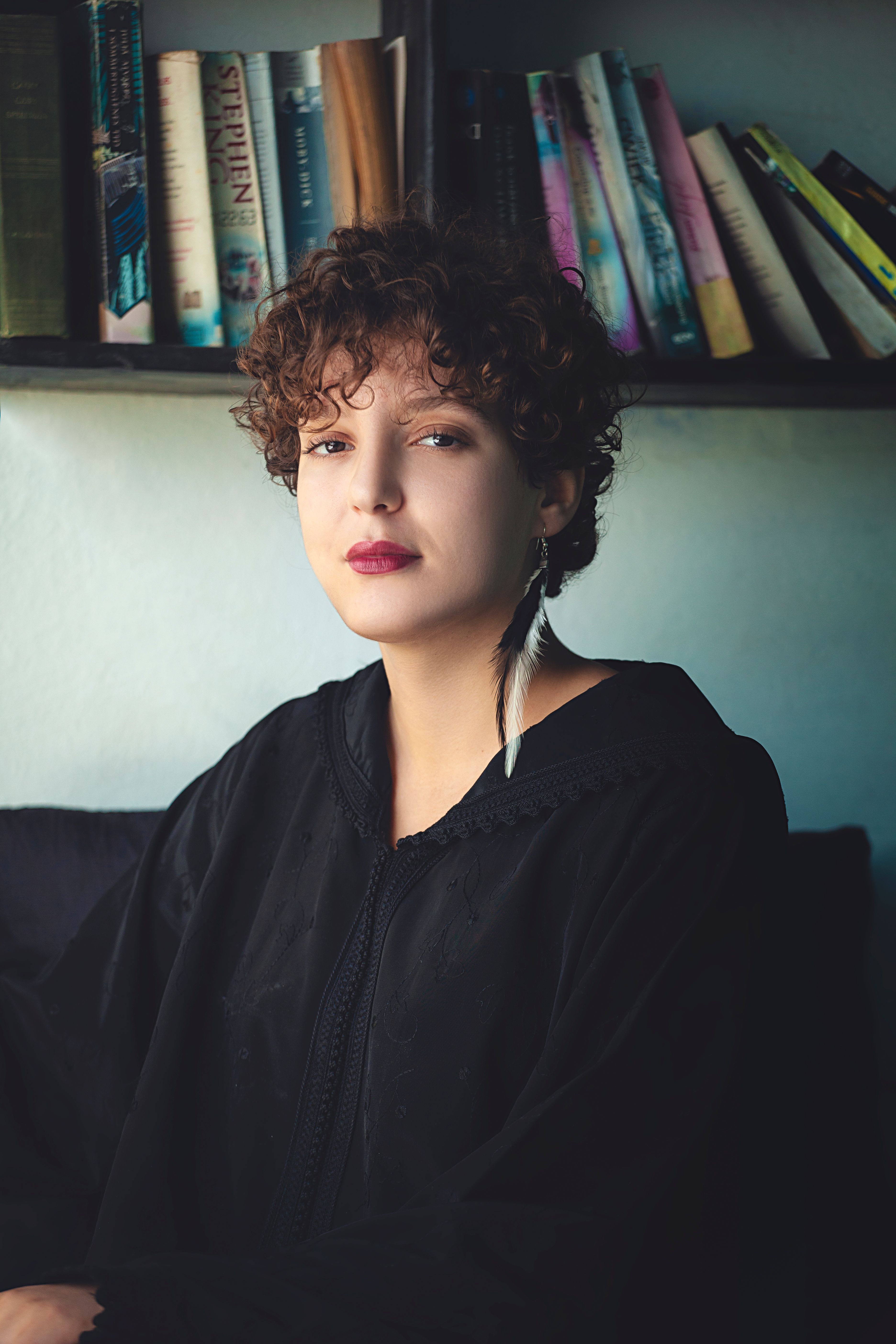 Stylish woman with curly hair and red lips, sitting by a bookshelf. Feather earring stands out.