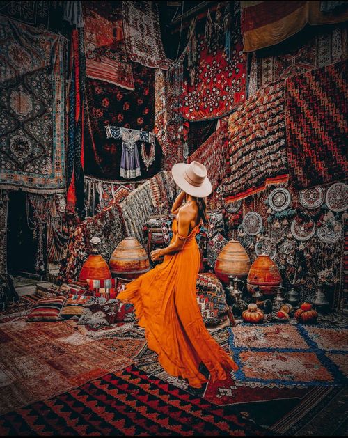 Free A woman in a flowing orange dress explores a vibrant market filled with ethnic fabrics and carpets. Stock Photo