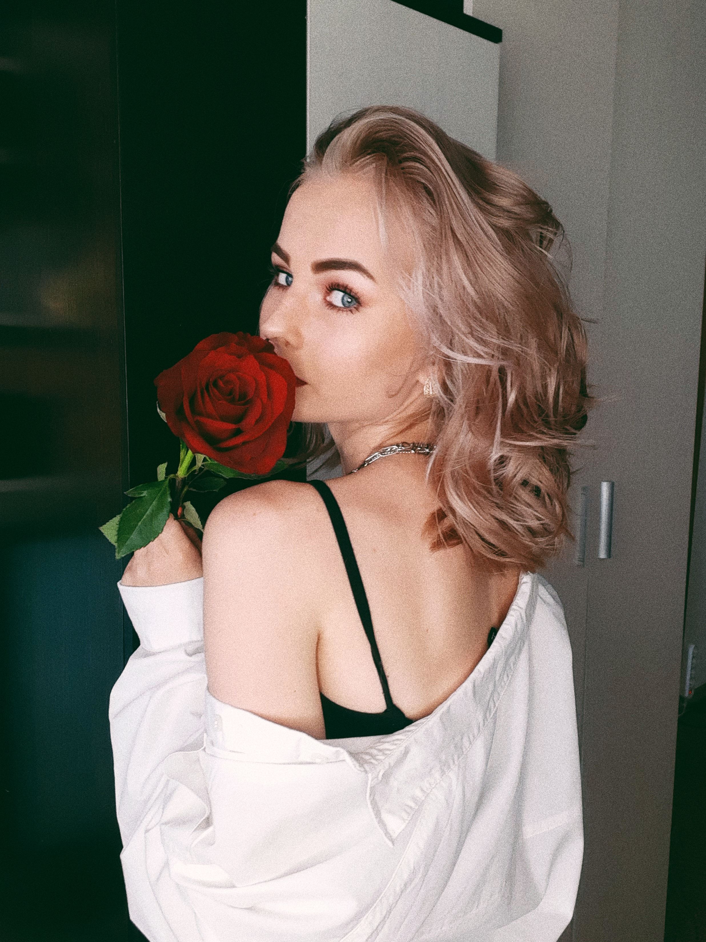 A woman with blonde hair holds a red rose, wearing a white shirt, looking over her shoulder.