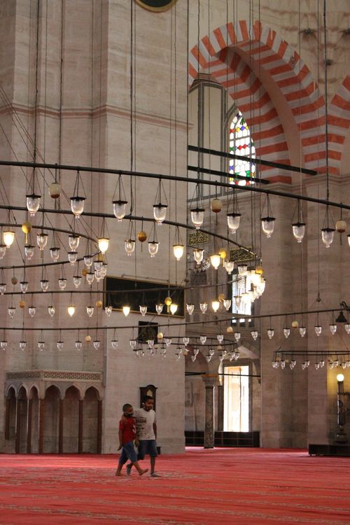 Free A grand mosque interior features arches, intricate chandeliers, and children walking on a carpet. Stock Photo