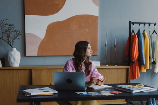 A woman at a stylish desk with clothing rack and abstract art, working in a modern workspace.