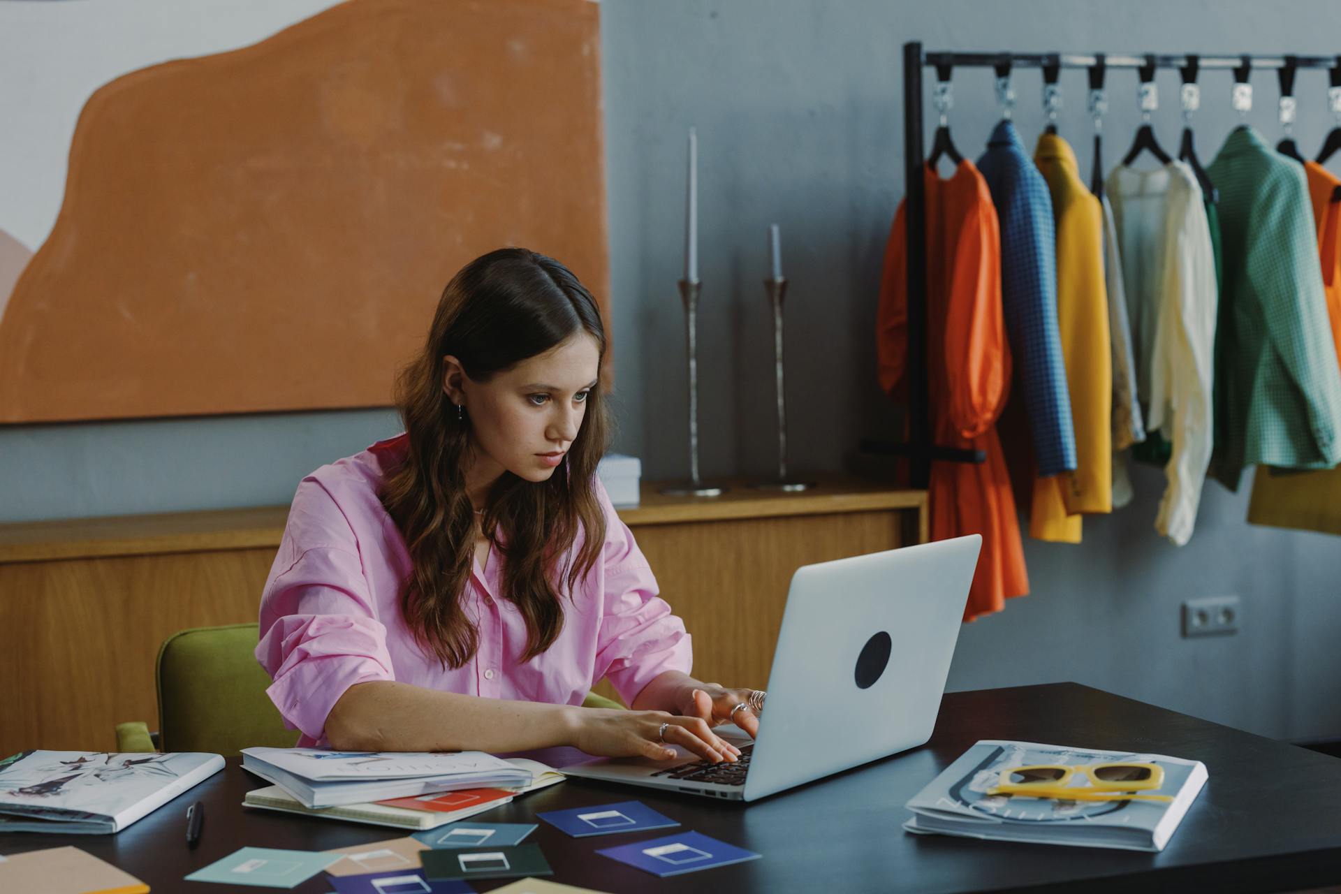 Businesswoman working at laptop in stylish office setting with clothes rack and design materials.