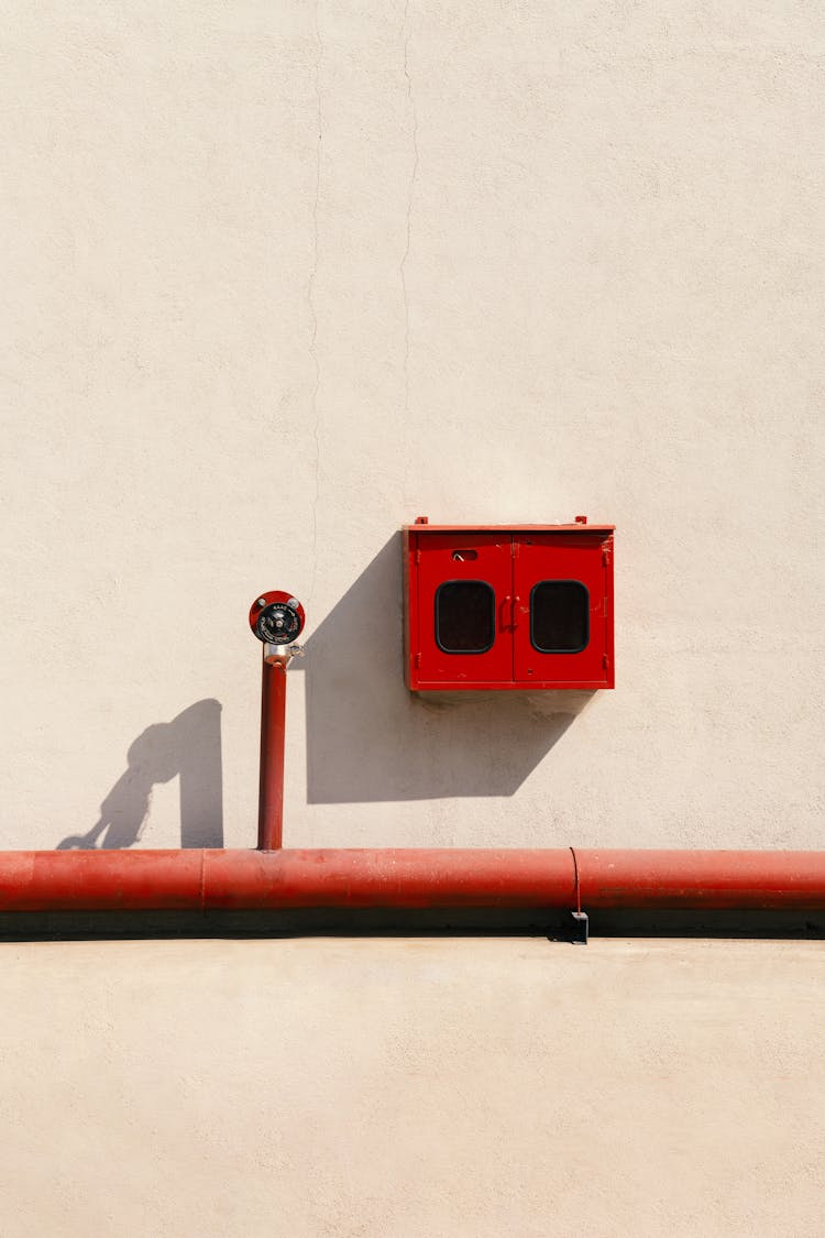 Abstract Image Of A Red Pipe And Electricity Box On A Beige Wall