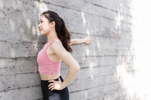 Asian woman stretching in pink top against a concrete wall. Outdoor fitness exercise.