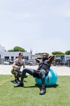 A man doing sit-ups on an exercise ball outdoors with a trainer guiding him, promoting fitness.