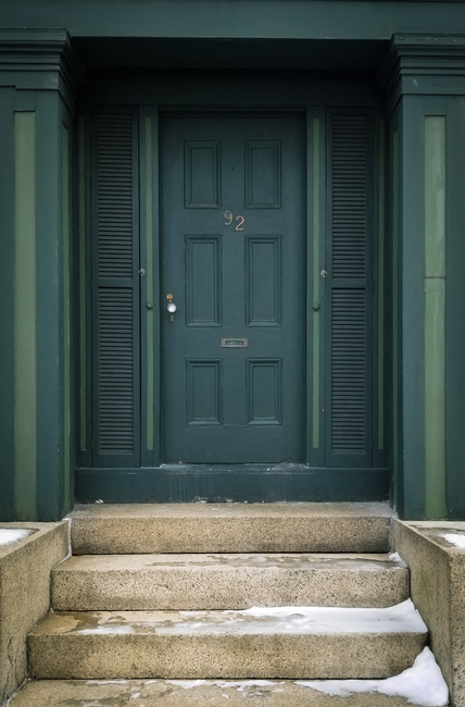 A vertical shot of a green door with concrete steps and light snow.