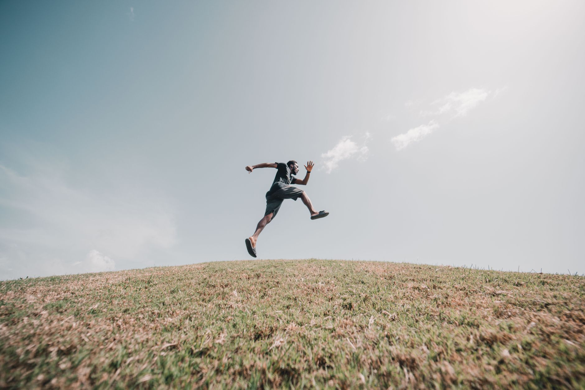 Man gracefully jumping on a grassy hill under a vast blue sky, showcasing energy and freedom.