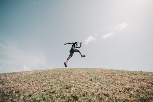 Man gracefully jumping on a grassy hill under a vast blue sky, showcasing energy and freedom.