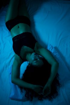 A serene woman lies on a bed in intimate black lingerie, illuminated by soft lighting.