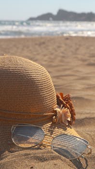 A straw hat and sunglasses rest on the sand with the ocean in the background, evoking a serene summer escape.