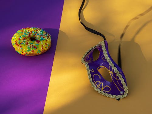 Close-Up Shot of a Carnival Mask beside a Donut on a Yellow Surface