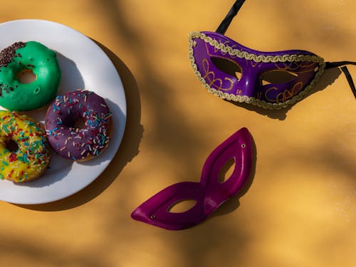 Close-Up Shot of Carnival Masks beside Donuts on a Yellow Surface