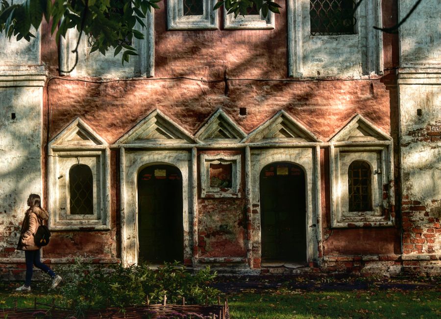 A historic monastery facade in Russia with sunlight casting shadows, a woman walking by.