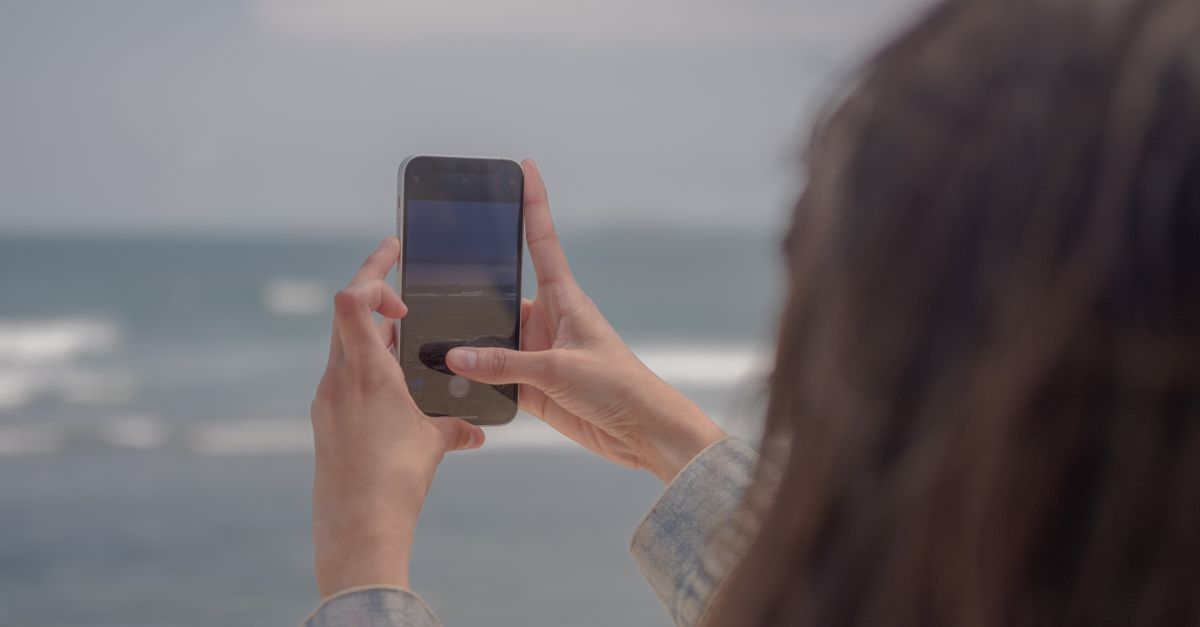 A woman captures a scenic beach view with her smartphone outdoors.