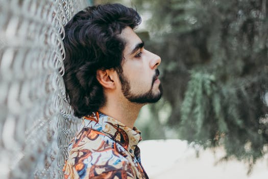 一张留着胡须的时尚年轻人靠在户外铁丝网栅栏上的侧面照片