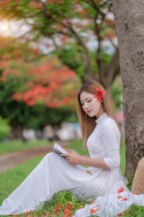 Free Woman in White Long Sleeve Dress Sitting on Green Grass while Holding a Book Stock Photo