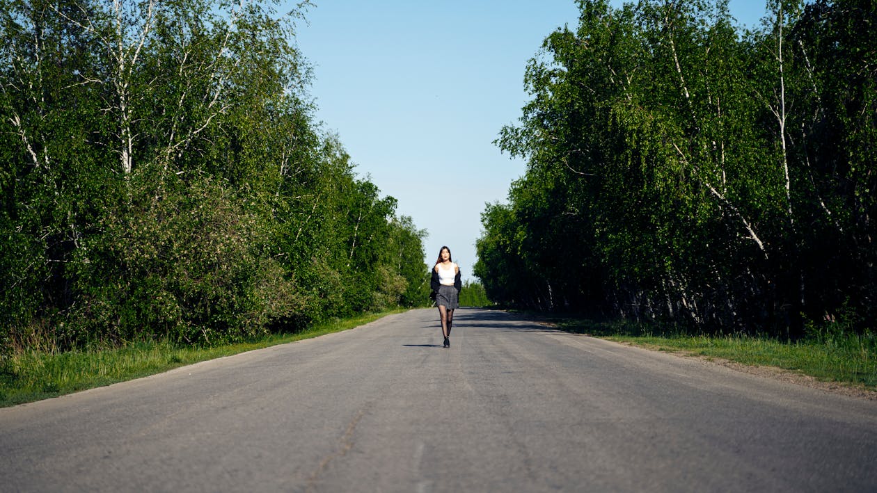 A Woman in White Tank Top Walking on the Road Between Green Trees