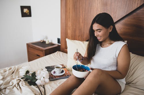 Free A Woman in White Tank Top Sitting on the Bed while Holding a Ceramic Bowl Stock Photo