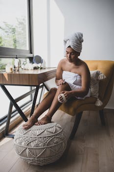 Woman wrapped in towel applies body scrub at home, practicing self-care.