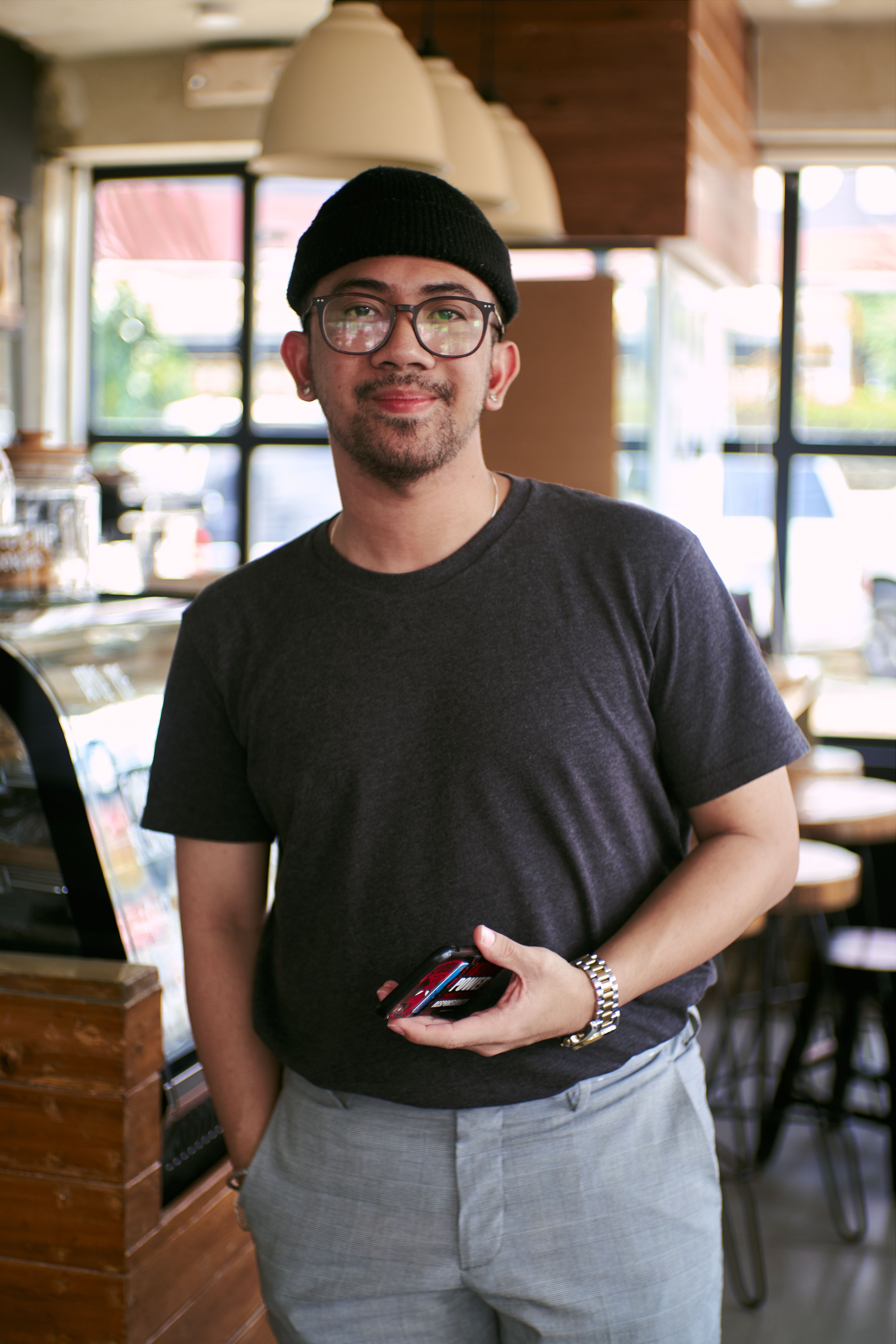 Fashionable Asian man with eyeglasses and beanie standing in a cafe, showcasing urban style.