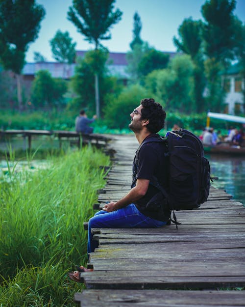 Free Side View of a Man with a Backpack Sitting on a Wooden Dock Stock Photo