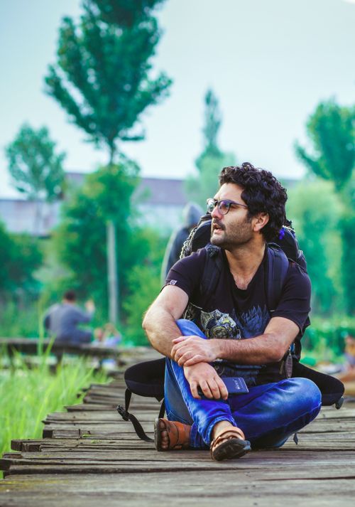 Free A man with a backpack and sunglasses sits on a wooden boardwalk, enjoying a scenic outdoor view. Stock Photo