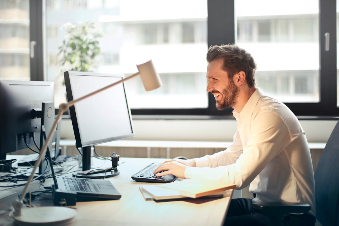 Man In Wit Overhemd Zittend Op Zwarte Rollende Stoel Terwijl Tegenover Zwarte Computerset En Glimlachen