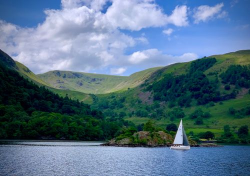 Free A sailboat glides on a tranquil lake surrounded by lush green mountains under a blue sky. Stock Photo
