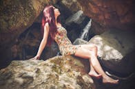 一位身着优雅花裙的女子，赤脚躺在瀑布边的岩石上，散发着大自然的宁静